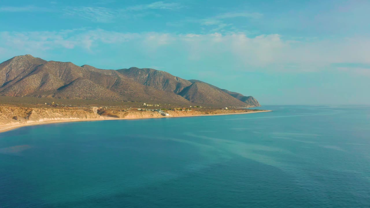 At 0:01, the El Sargento La Ventana mountain ridge remains centered over calm blue water, with a narrow tan shoreline and no visible people.