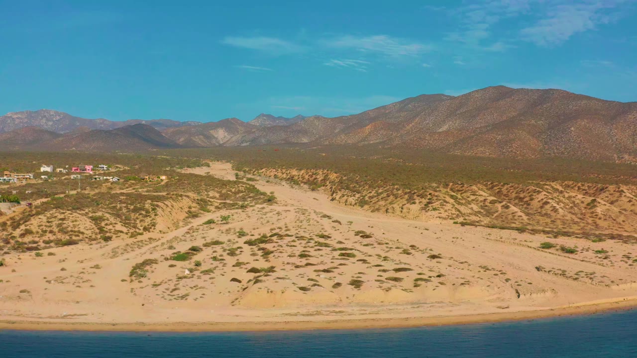 Aerial view in El Sargento La Ventana, Mexico, showing the beach edge lower in frame, scrub-covered desert, a small cluster of distant structures, mountains, and no visible people.
