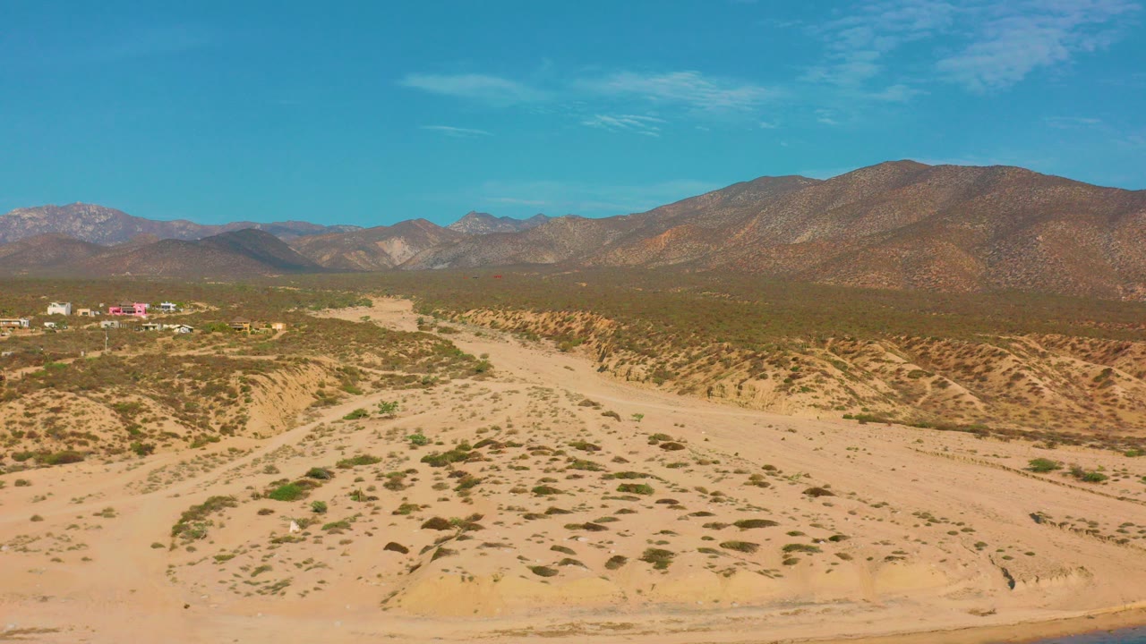 Opening aerial frame over El Sargento La Ventana, Mexico, showing a sandy wash, scattered desert shrubs, distant small buildings at left, mountain ridges, blue sky, and no visible people.