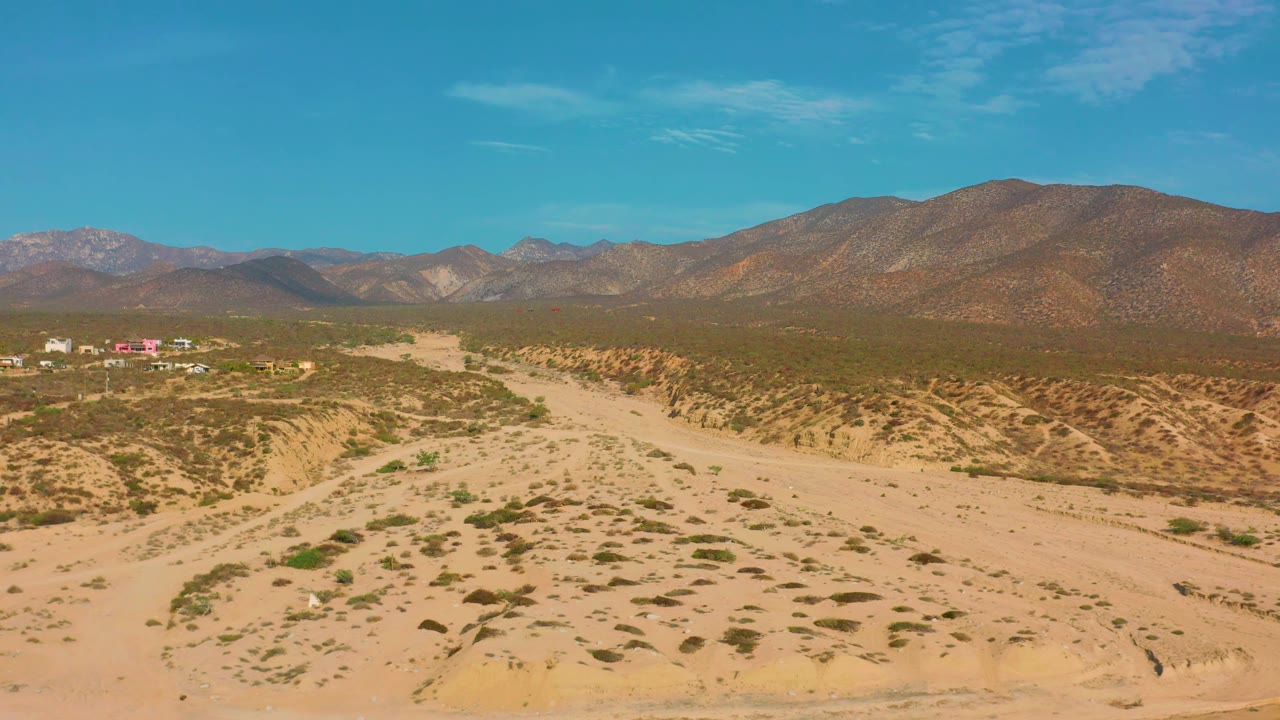 Mid-clip aerial view over El Sargento La Ventana, Mexico, showing textured sandy terrain, scattered green shrubs, a small distant settlement on the left, mountain horizon, and no visible people.