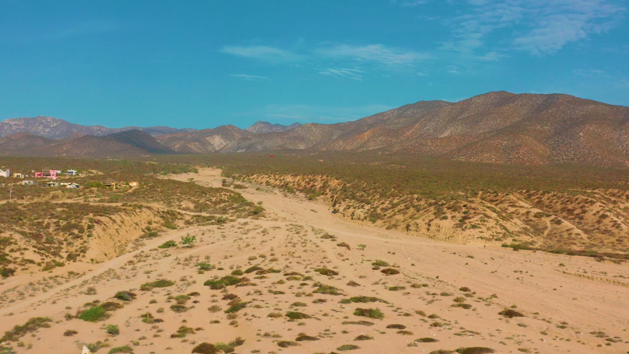Opening aerial frame over El Sargento La Ventana, Mexico, showing a pale sandy wash, scattered desert brush, distant mountains, small structures at far left, and no visible people.