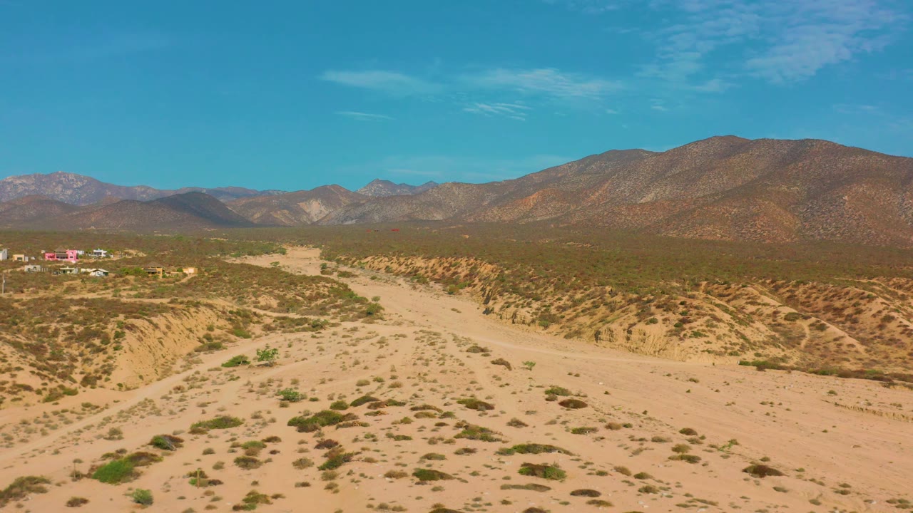 Aerial frame from El Sargento La Ventana, Mexico, showing pale desert sand in the foreground, low green brush, ridged hills, distant mountains, and no visible people.