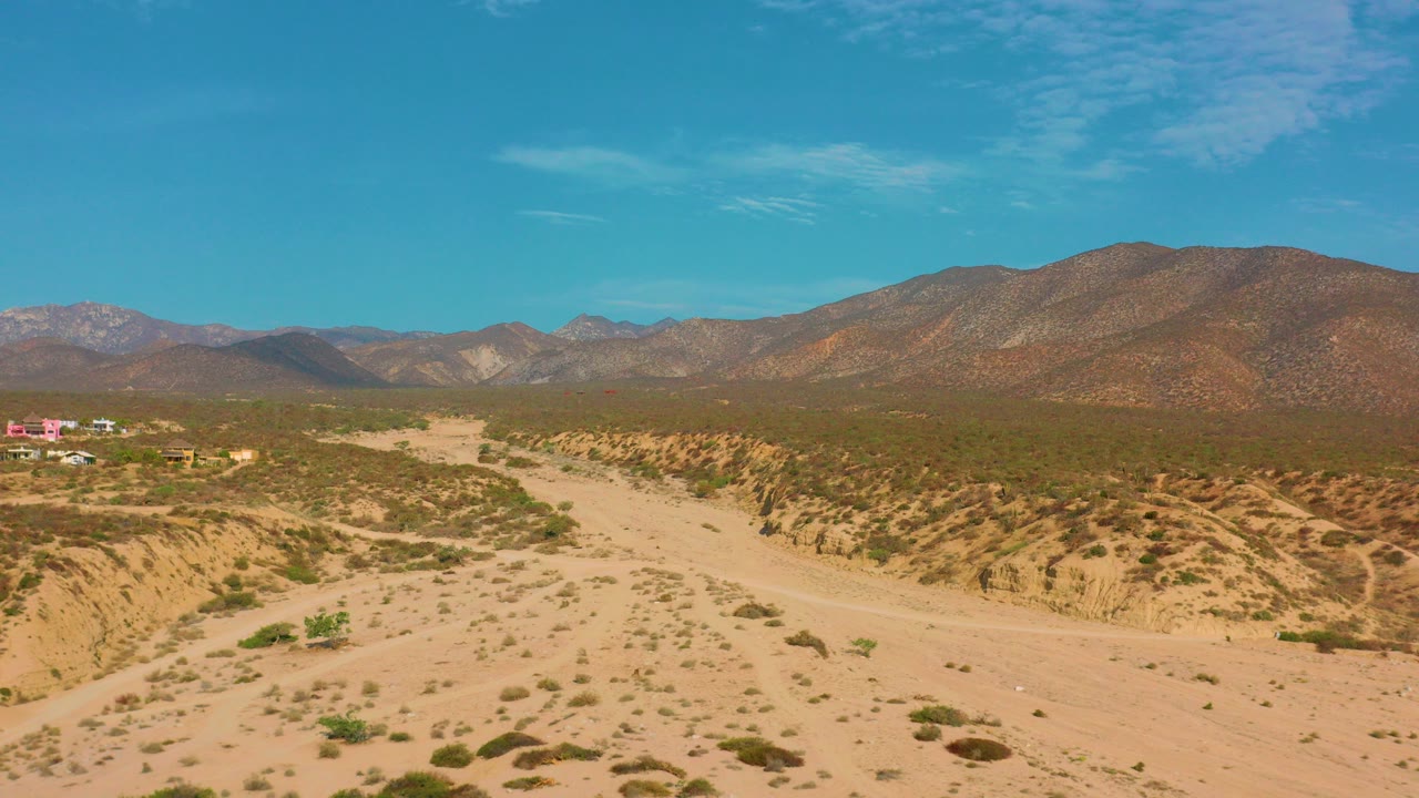 Opening frame of an aerial view over El Sargento La Ventana, Mexico, with a pale desert road, scrub-covered wash, distant buildings at left, mountains on the horizon, and no visible people.
