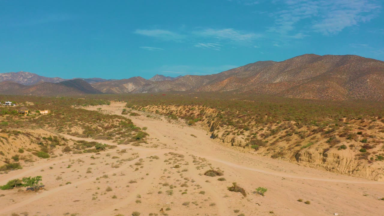 A wide aerial frame shows sandy desert washes, sparse green scrub, distant mountains, and blue sky near El Sargento La Ventana, Mexico, with no visible people.
