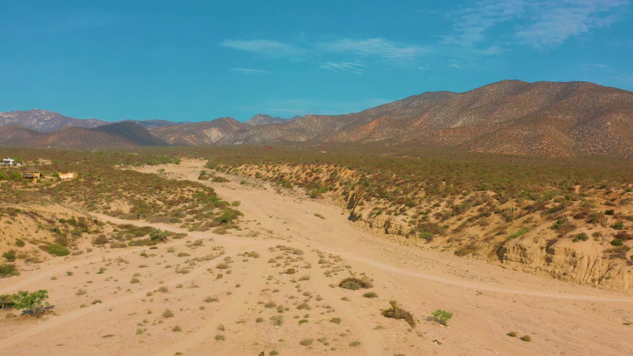 An elevated view shows eroded desert banks crossing the right side of the frame, open sand below, and mountains stretching across the horizon near El Sargento La Ventana, Mexico, with no visible people.