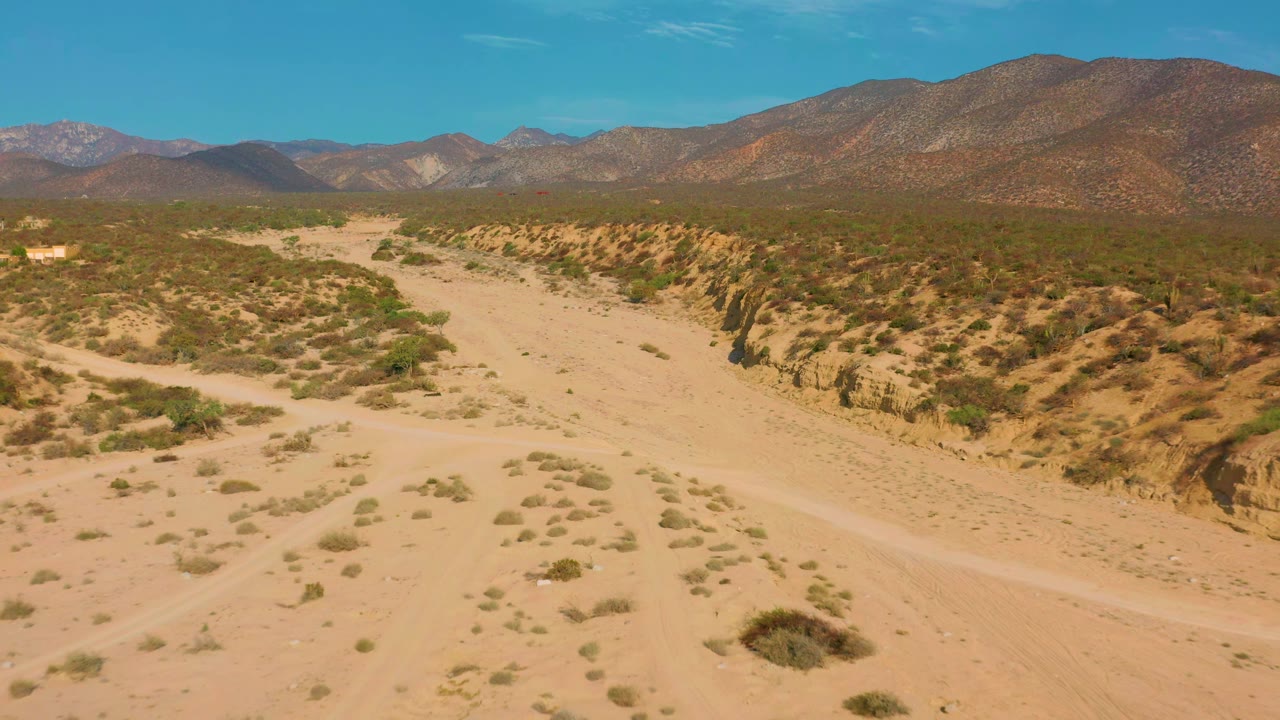 A wide sandy track runs through scrubby dry ground in El Sargento La Ventana, with a raised bank on the right, mountains beyond, and no visible people.
