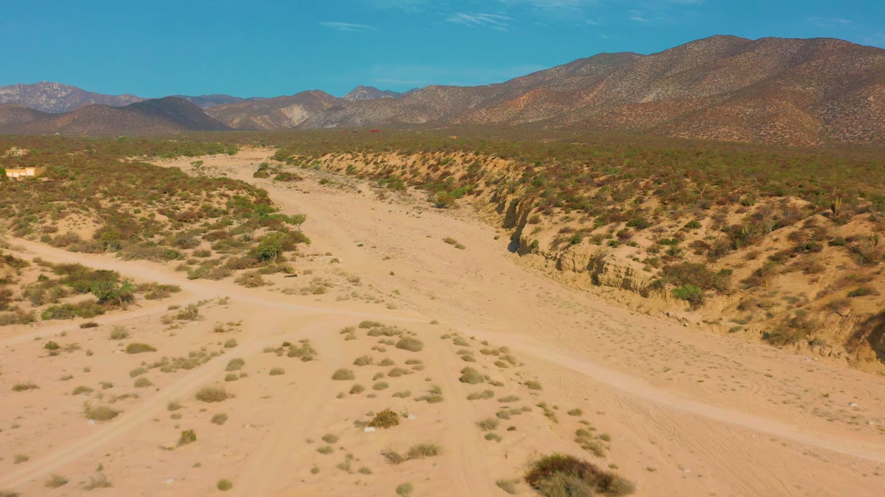 The sandy roadway and wash sit between low shrub-covered ridges in El Sargento La Ventana, with brown mountains across the horizon and no visible people.