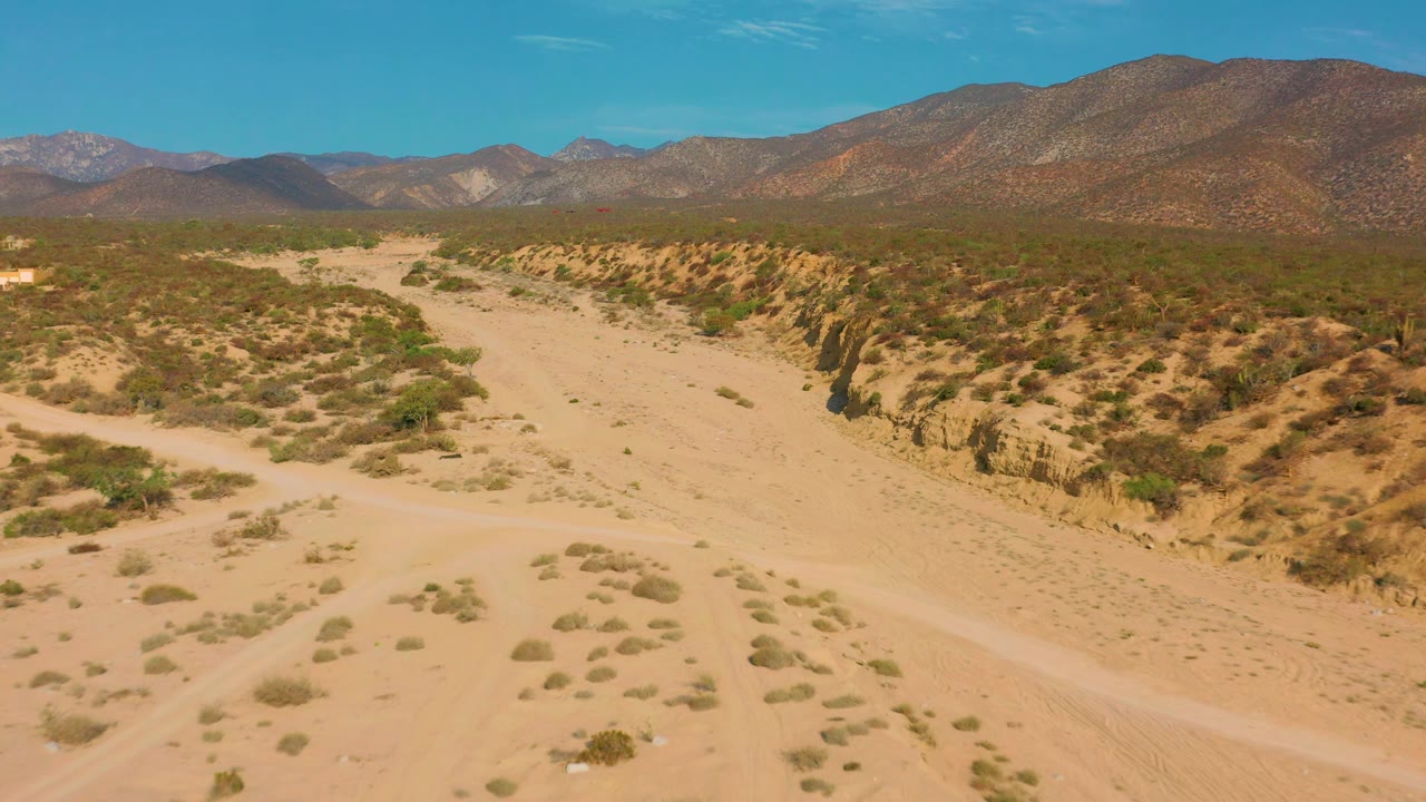 A pale sand road crosses the foreground of El Sargento La Ventana terrain, with eroded banks, scattered shrubs, distant mountains, and no visible people.