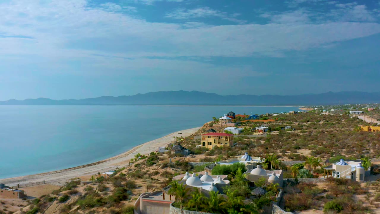 Elevated view over El Sargento La Ventana, Mexico, showing blue-green water, a pale curving beach, dry coastal lots, scattered homes, distant mountains, and no visible people.
