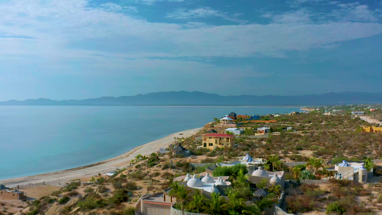 Aerial shoreline view with calm water, beach, scrubland, and houses near the coast.