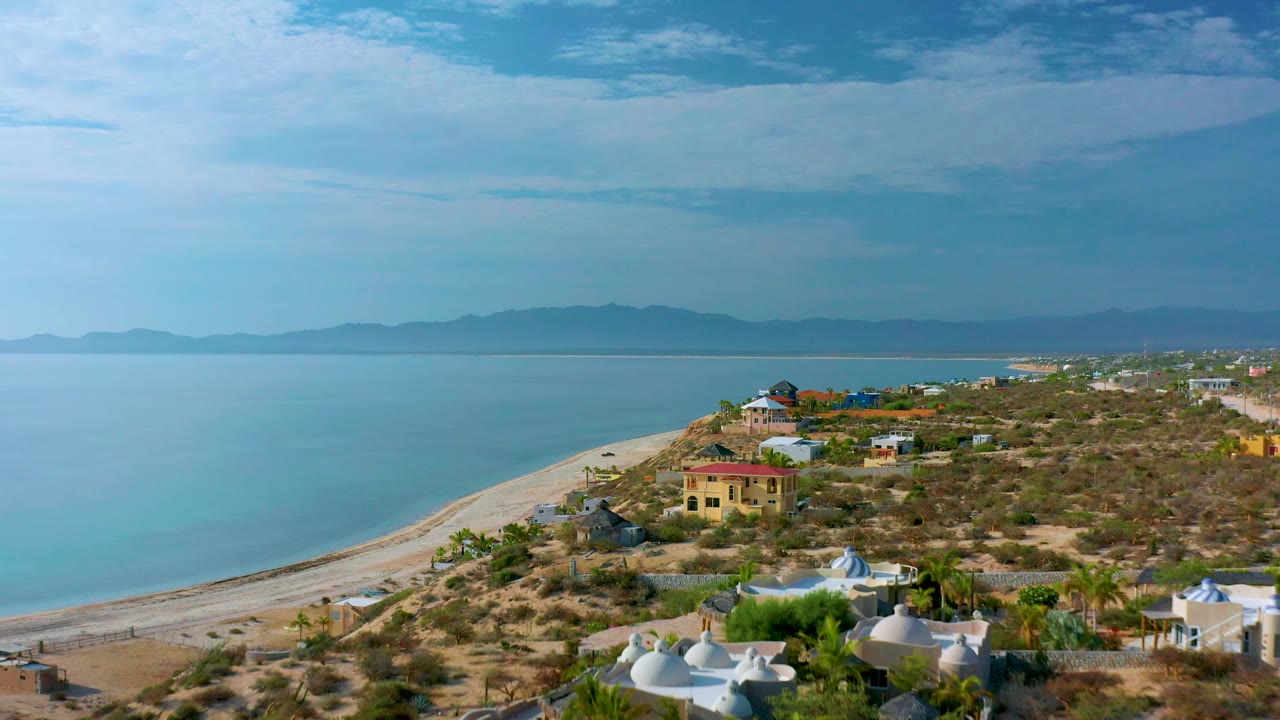 El Sargento La Ventana, Mexico, appears from an elevated coastal angle with a broad water surface, narrow sandy shore, residential buildings, dry vegetation, distant mountains, and no visible people.