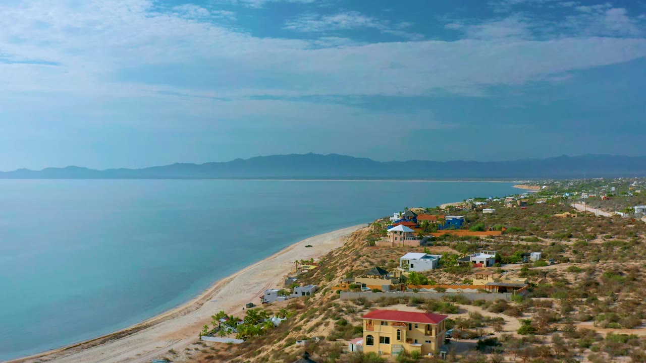 Coastal homes and scrubland beside a curved beach and calm water.