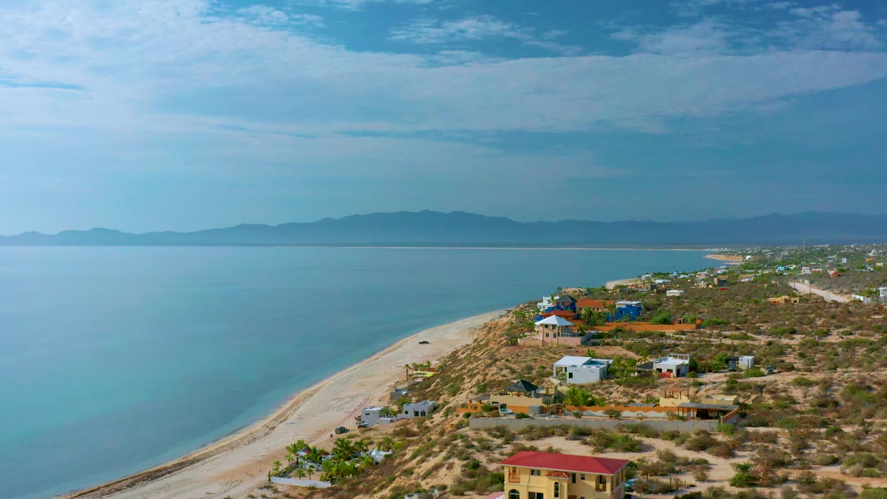 Third frame of El Sargento La Ventana, Mexico with more homes visible along the right side, pale sand below, broad blue water to the left, and no visible people.