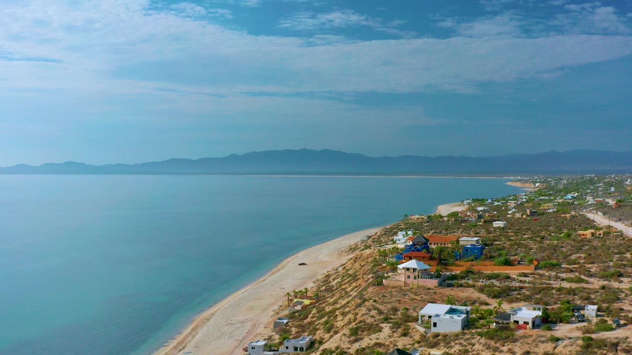Opening aerial view over El Sargento La Ventana with turquoise water on the left, a pale beach curving past scrubland and scattered houses, distant mountains, and no visible people.