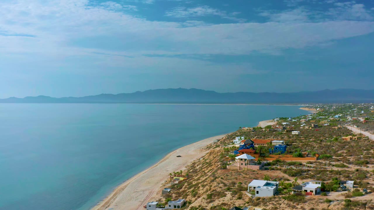 Early drone frame over El Sargento La Ventana showing a long sandy shoreline, blue-green sea, low houses among shrubs, a mountain range across the water, and no visible people.
