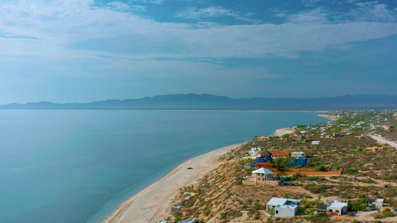 Aerial coastal frame in El Sargento La Ventana with the shoreline running from lower left toward the settlement, desert lots and houses to the right, mountains on the horizon, and no visible people.