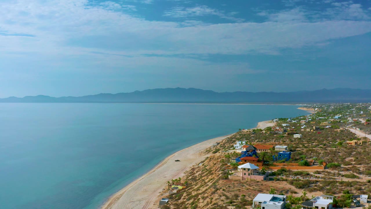 Wide aerial frame of calm sea, sandy beach, shrubs, houses, and distant mountains.