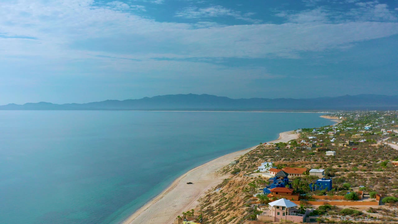 Opening aerial frame over El Sargento La Ventana, Mexico, with turquoise sea on the left, pale beach curving through the center, coastal houses on the right, distant mountains, and no visible people.
