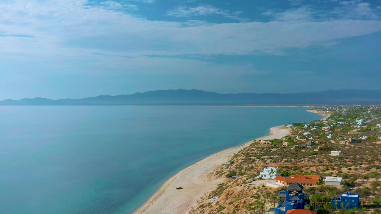 Aerial frame over El Sargento La Ventana, Mexico, showing the beach curve more centrally, with turquoise water to the left, clustered homes to the right, mountains on the horizon, and no visible people.