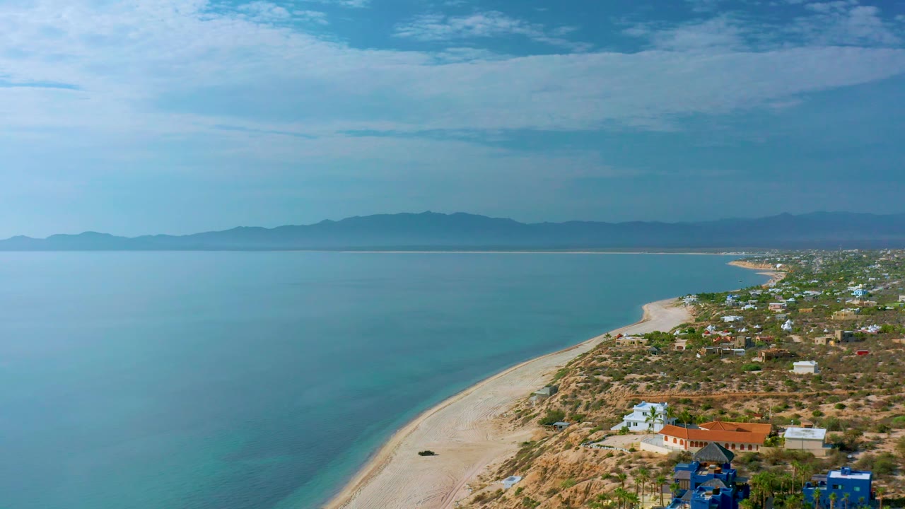Aerial view in El Sargento La Ventana, Mexico, with a pale beach running diagonally through the frame, calm sea filling the left side, homes along the right edge, distant mountains, and no visible people.
