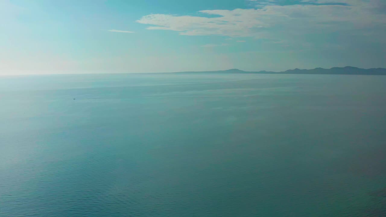 Wide aerial view over blue ocean at El Sargento La Ventana, Mexico, with pale glare on the water, distant low land along the horizon, thin clouds above, and no visible people.