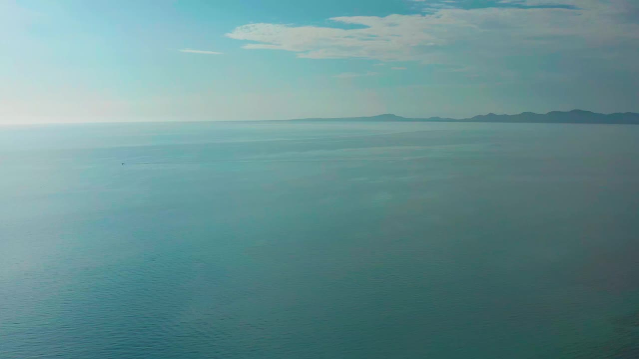 Aerial ocean frame from El Sargento La Ventana, Mexico, showing a bright sheen across the blue water, distant dark landforms near the horizon, light cloud patches, and no visible people.