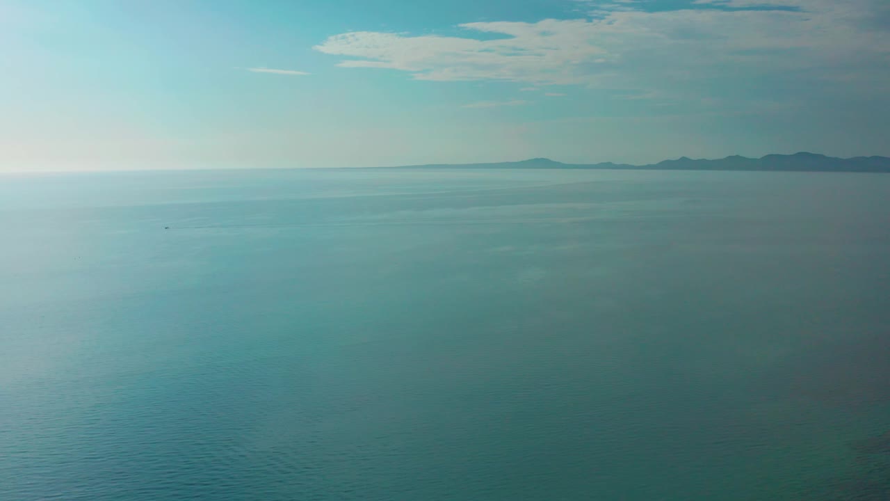 High view over turquoise and deep blue ocean at El Sargento La Ventana, Mexico, with open water dominating the frame, far shoreline shapes at the upper edge, scattered cloud cover, and no visible people.