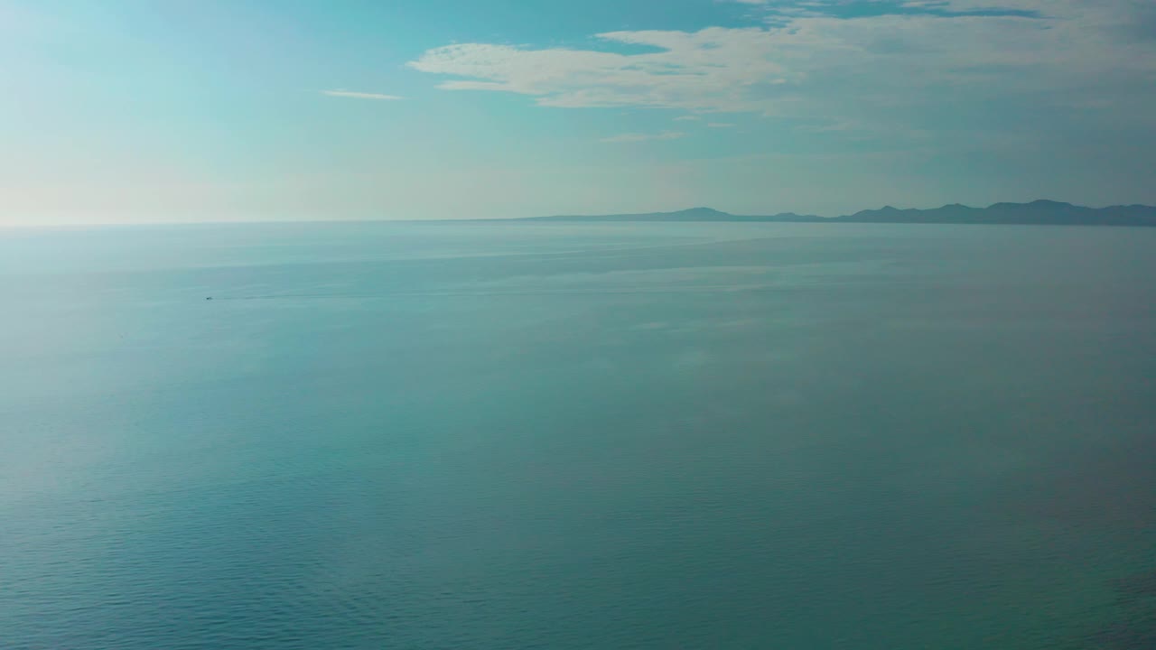 Aerial view at El Sargento La Ventana, Mexico, with blue-green ocean filling the frame, distant land on the horizon, a tiny dark speck on the water far from camera, cloud bands above, and no visible people.