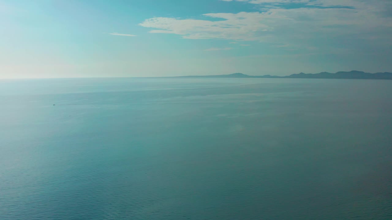 Frame at 0:02 keeps the El Sargento La Ventana horizon high in view, with turquoise ocean below, low island shapes in the distance, thin cloud cover, and no visible people.