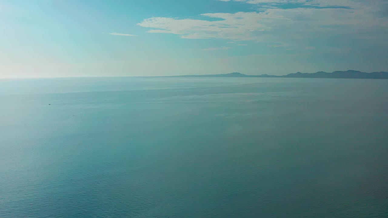 Mid-clip frame shows a broad blue ocean expanse near El Sargento La Ventana, with the distant mountainous coastline along the upper right, pale haze at the horizon, and no visible people.