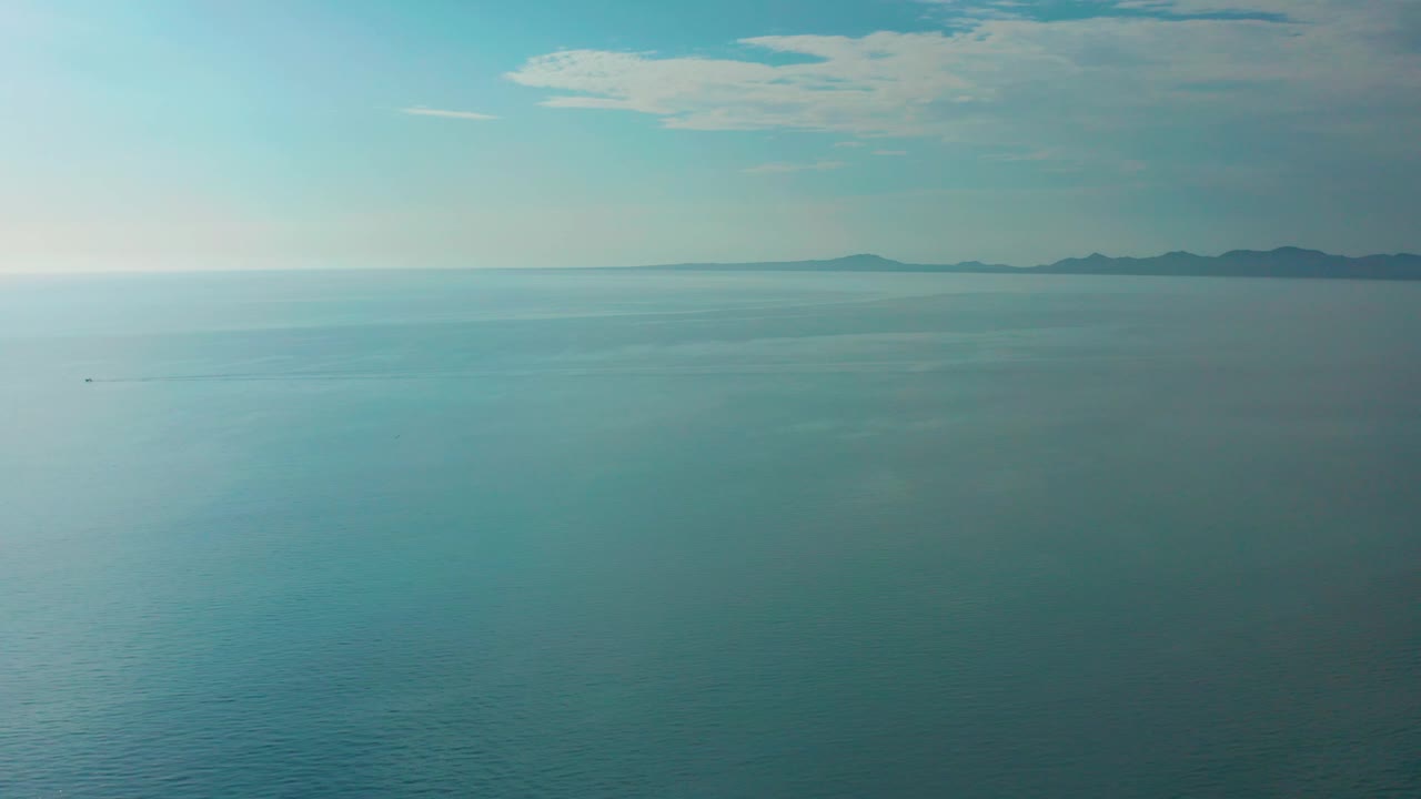 Fourth frame holds on the El Sargento La Ventana seascape, with calm ocean foreground, hazy coastline stretching across the background, thin clouds overhead, and no visible people.