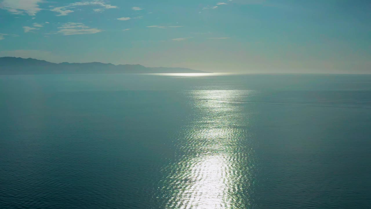 High aerial frame from El Sargento La Ventana, Mexico, showing rippled teal water, a wide silver glare path near the lower middle, low mountains across the left horizon, and no visible people.