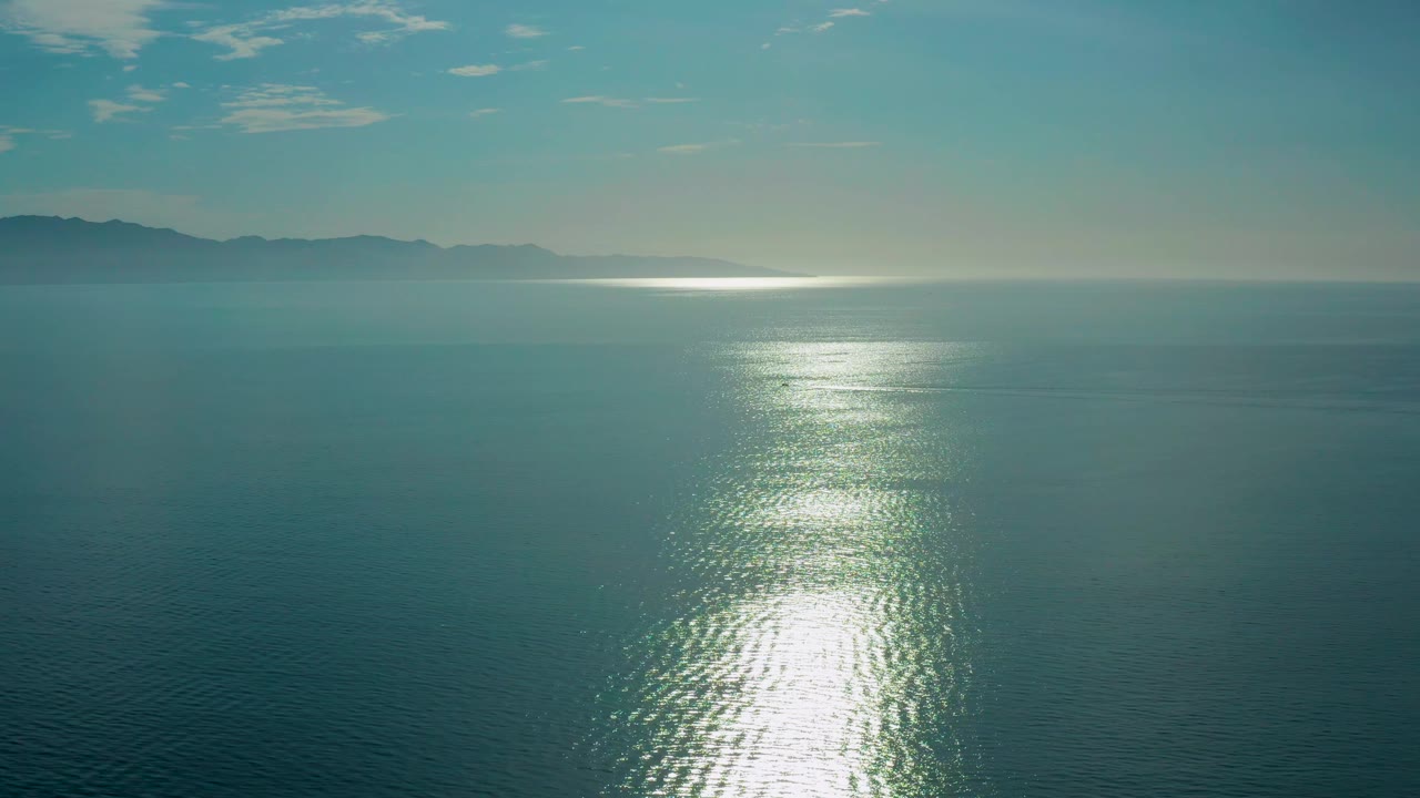 Open sea in El Sargento La Ventana, Mexico, with the bright sun reflection centered lower in frame, muted blue-green water around it, hazy landforms to the left, and no visible people.