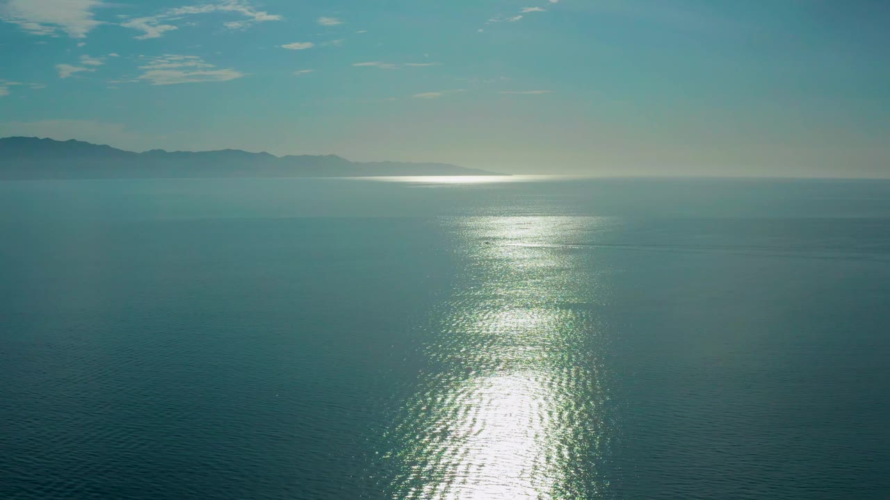 Wide aerial view in El Sargento La Ventana, Mexico, where teal water fills most of the frame, bright glare breaks across the lower water, mountains sit low on the left horizon, and no visible people appear.