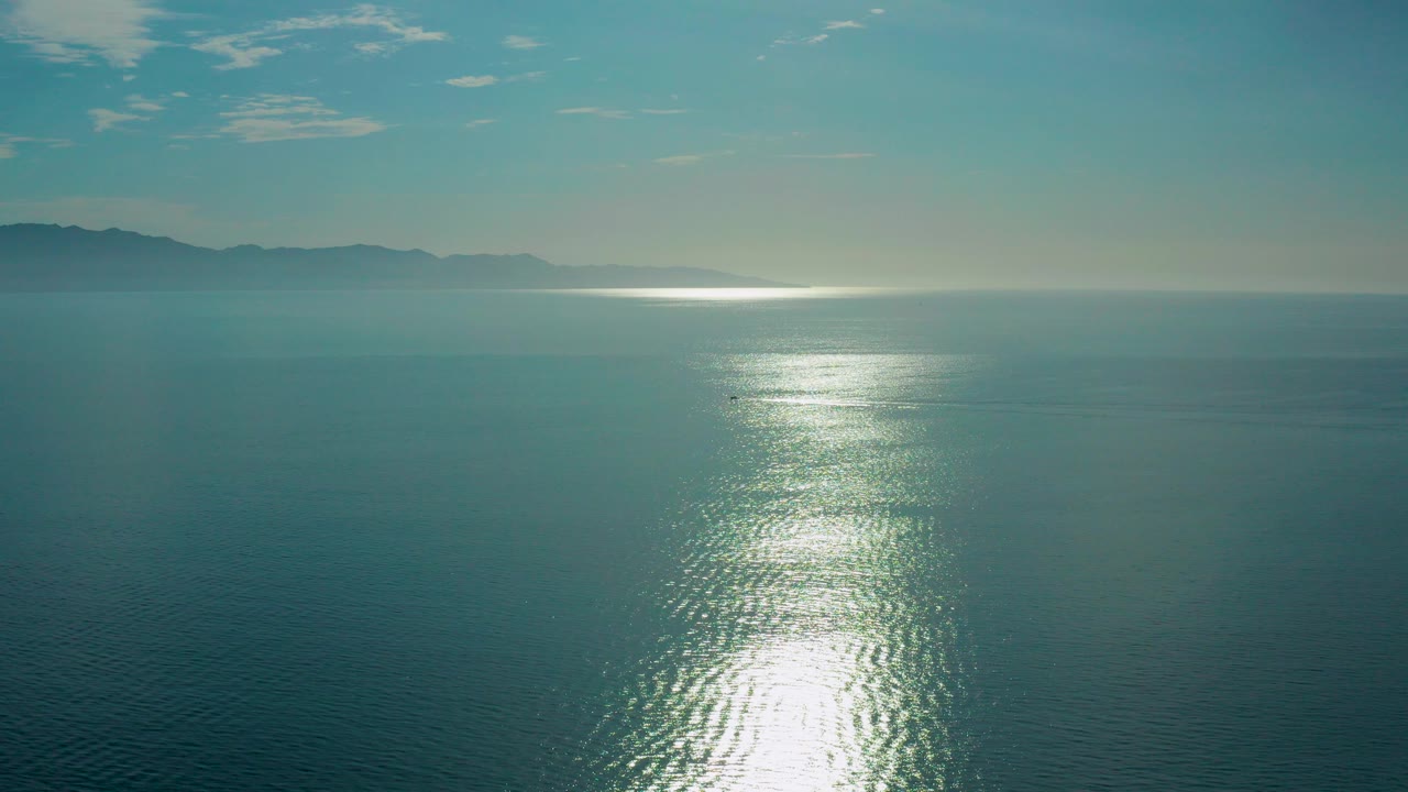 Opening frame over blue-green ocean in El Sargento La Ventana, Mexico, with a bright sun reflection on rippled water, distant mountains on the horizon, and no visible people.