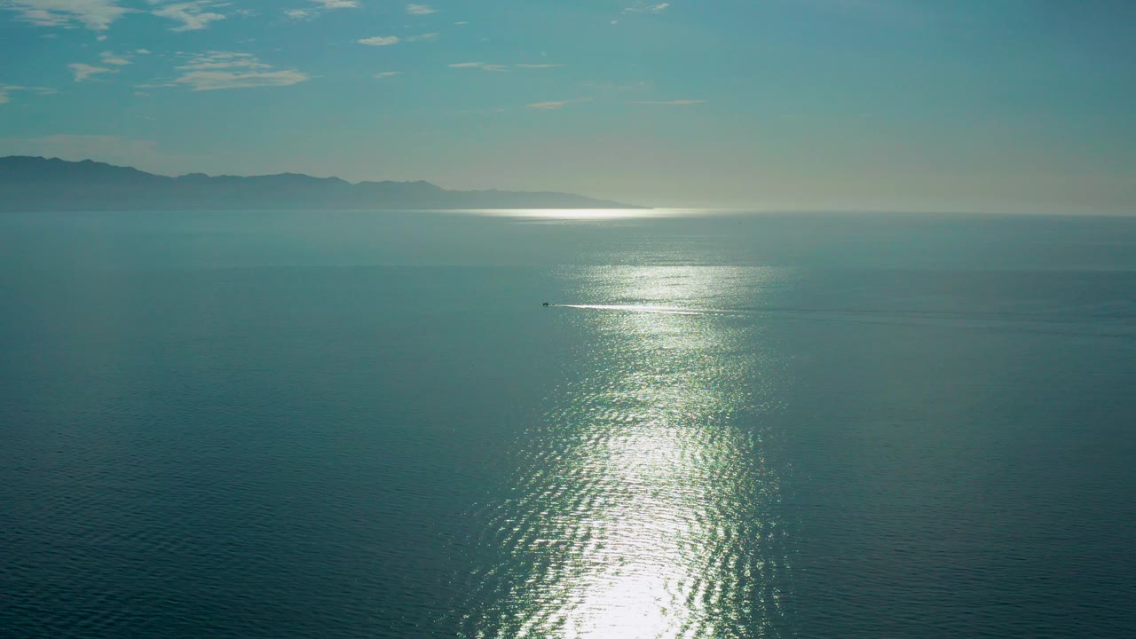 Wide aerial view over blue-green ocean in El Sargento La Ventana, Mexico, with bright sun glare on ripples, hazy mountains on the horizon, a small distant watercraft, and no visible people.