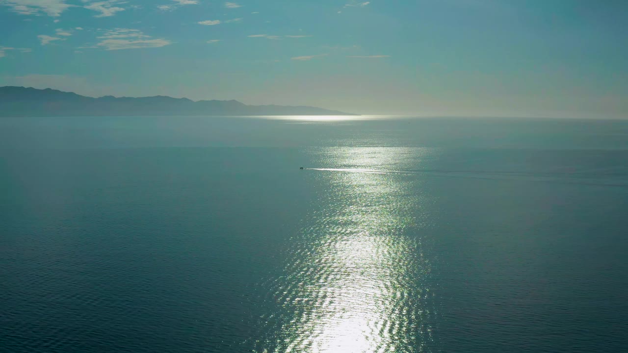High ocean view in El Sargento La Ventana, Mexico, where a small watercraft sits left of the sunlit water path below a hazy coastline, with no visible people.