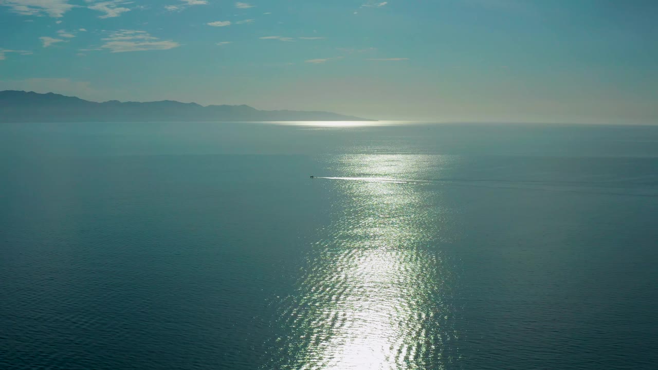Aerial ocean frame from El Sargento La Ventana, Mexico, with the sun reflection centered on textured water, low mountains in haze, a tiny distant craft, and no visible people.