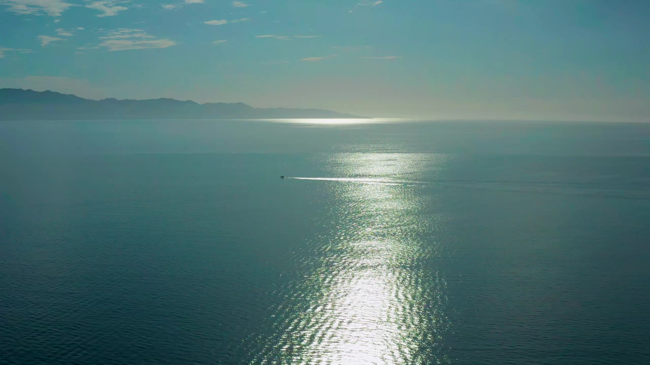 The ocean surface at El Sargento La Ventana, Mexico is crossed by a vertical strip of bright sunrise reflection, with hazy mountains along the far horizon and no visible people.