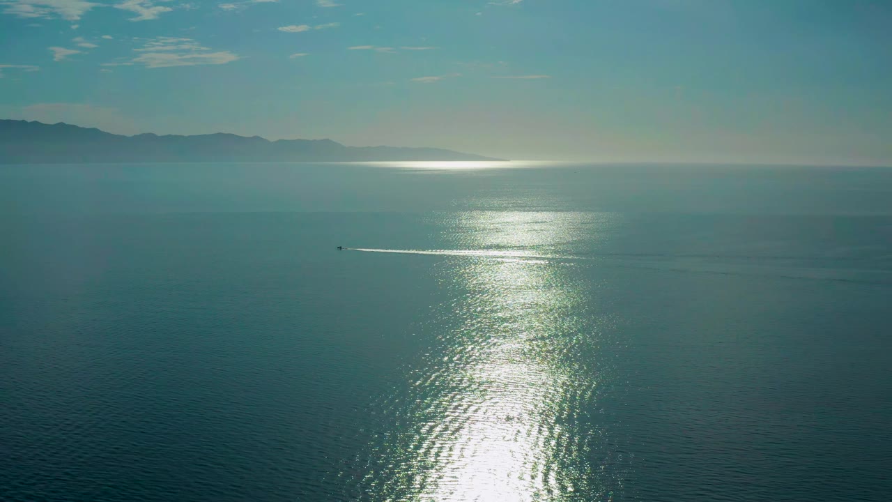 A small boat and narrow white wake cross the midground below the bright water reflection in El Sargento La Ventana, with mountains faint on the horizon and no visible people.