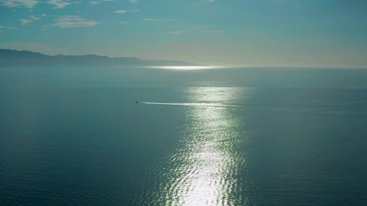 The sun glare spreads down the center of the ocean while a small boat sits left of the light path, with the El Sargento La Ventana mountain horizon visible and no visible people.