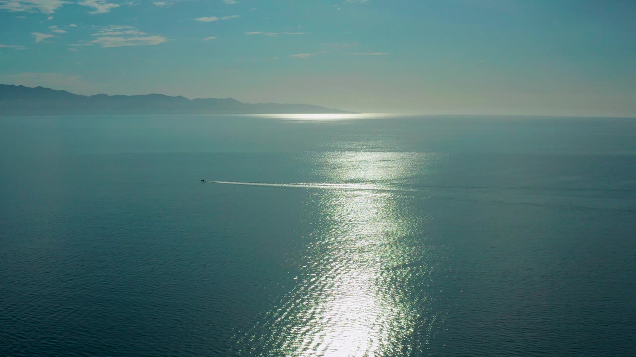 At the start of the clip, a wide aerial view over El Sargento La Ventana shows dark teal ocean, bright sun glare on the water, distant mountains, one small boat left of center, and no visible people.
