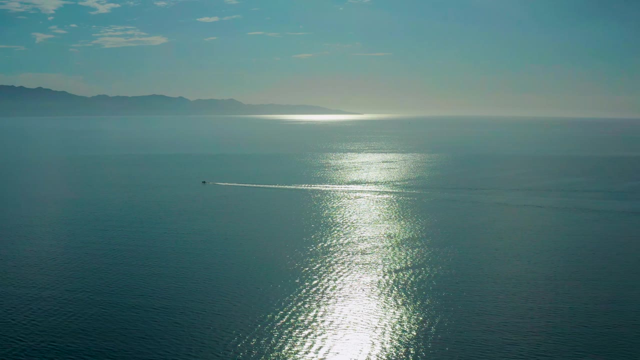 One second in, the boat sits slightly farther across the dark water, leaving a thin white wake below the bright sun path, with El Sargento La Ventana mountains on the horizon and no visible people.