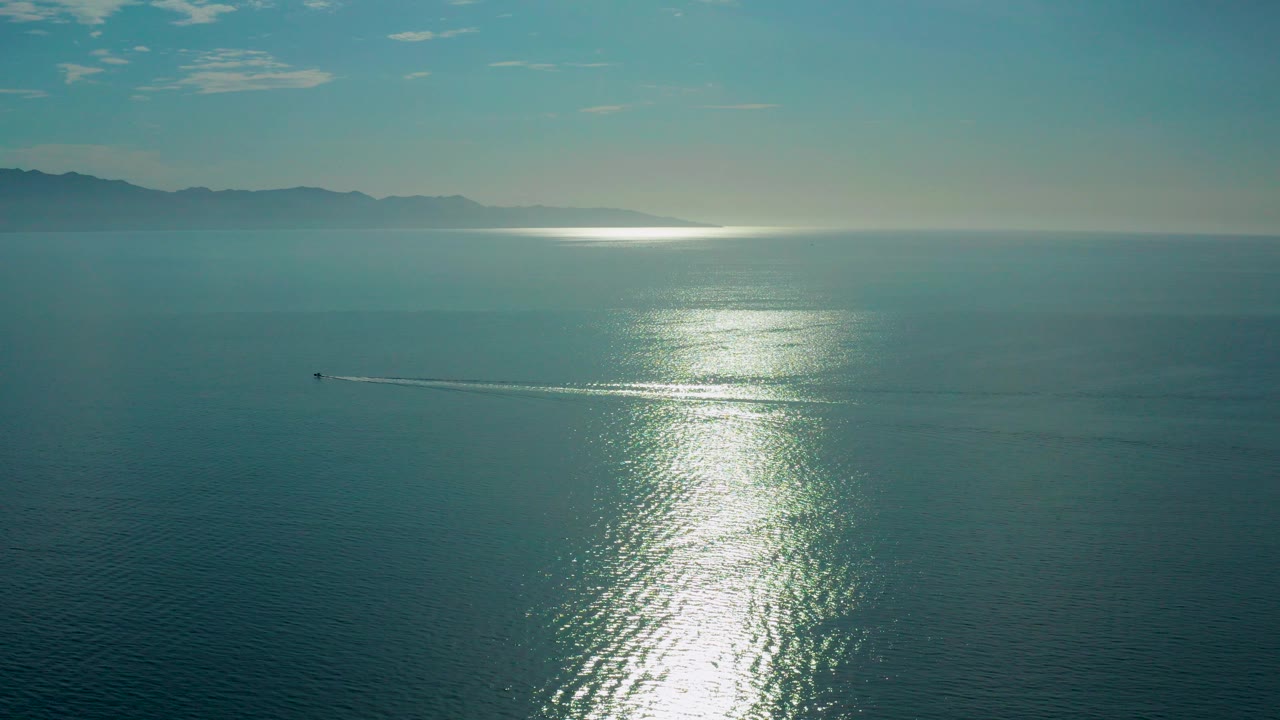 Aerial view over teal water in El Sargento La Ventana, Mexico, with a bright sun reflection near center, hazy mountains on the horizon, a small boat at left, and no visible people.