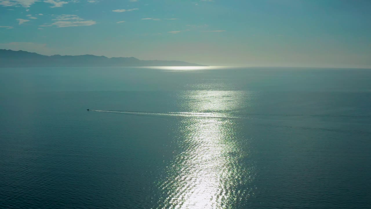 Wide aerial frame of El Sargento La Ventana water with the sun reflection stretching down the sea, a small boat farther left of the glare, distant mountains, and no visible people.