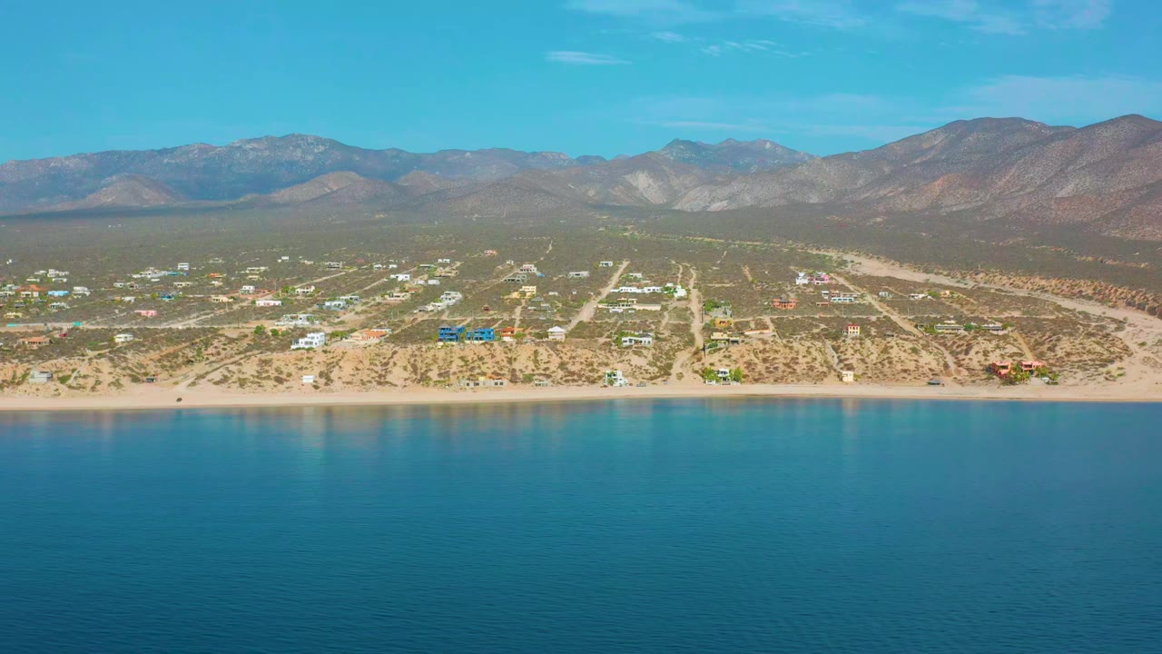 Aerial coastal view with calm blue water, beach, roads, buildings, and mountain ridge.