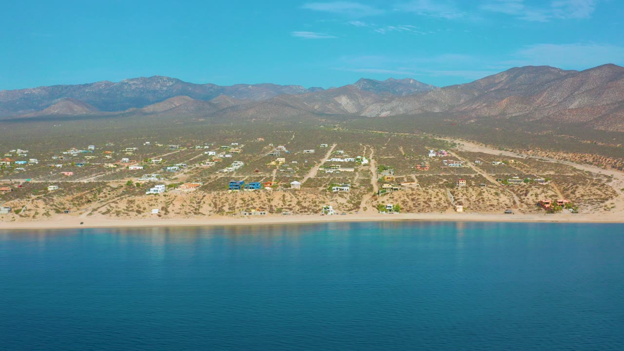 Opening frame of El Sargento La Ventana, Mexico, with deep blue water in front, a thin sandy beach, scattered coastal houses, dry terrain, mountains on the horizon, and no visible people.