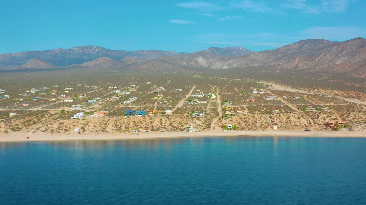 Aerial view over El Sargento La Ventana, Mexico, with blue water in front, a narrow beach, scattered coastal homes, desert terrain, distant mountains, and no visible people.