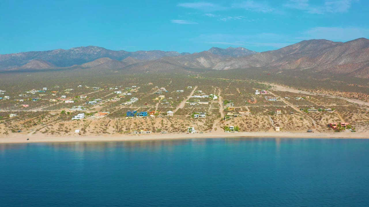 Wide aerial frame in El Sargento La Ventana, Mexico, showing turquoise water, a pale shoreline, small houses and roads inland, mountain ridges behind them, and no visible people.