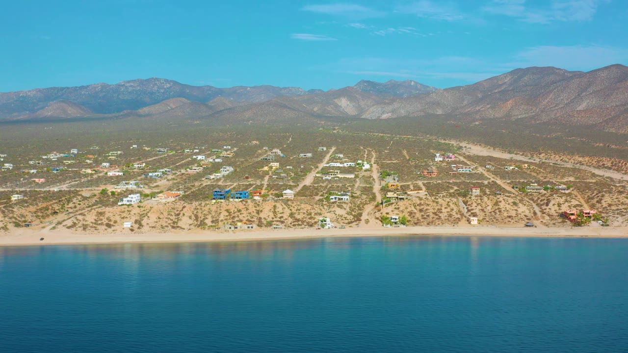 Aerial coastline view of El Sargento La Ventana, Mexico, with calm blue sea below, a thin beach band, scattered buildings across a dry plain, mountains on the horizon, and no visible people.